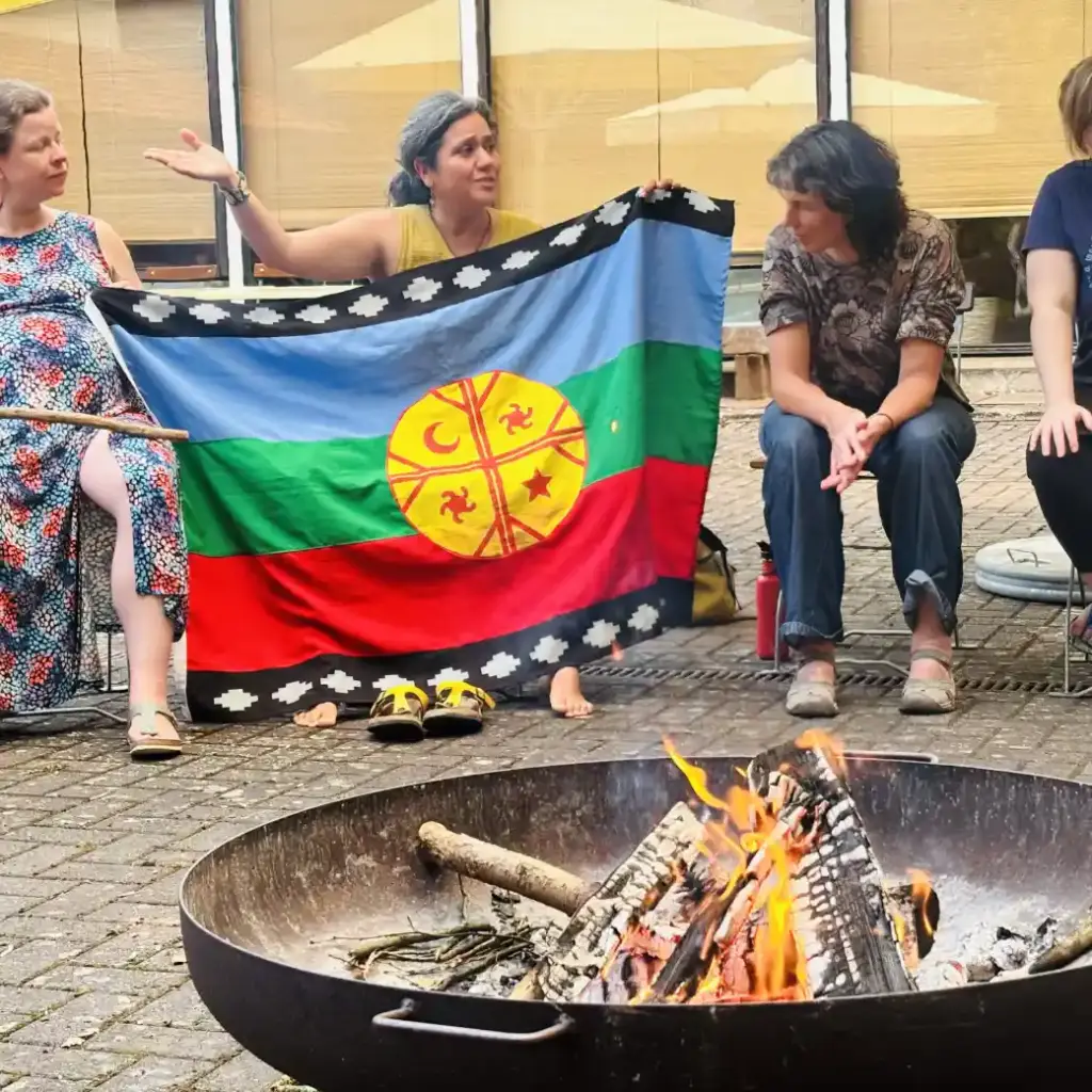 Fransisca Gallegos zeigt die Flagge der Mapuche und erklärt ihre Bedeutung. Vor ihr eine Feuerschale mit Feuer darin.